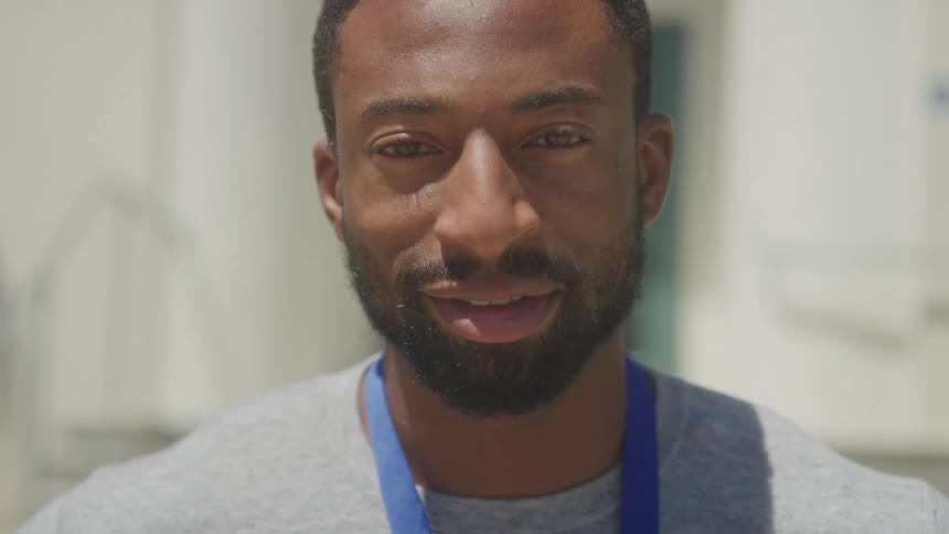 A young adult man with a beard and short hair wearing a grey shirt and a blue lanyard looks to his right then turns to face the camera and smiles with a light colored building in the blurred background