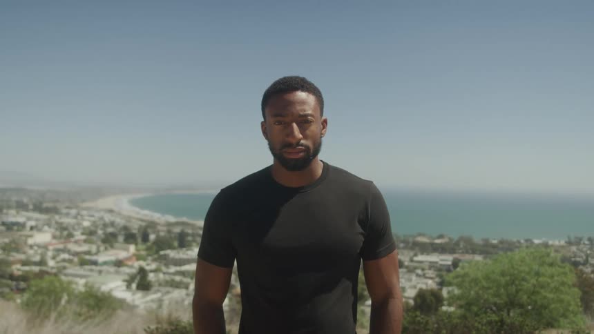 A man with a beard stands on a cliff at midday wearing a black tshirt