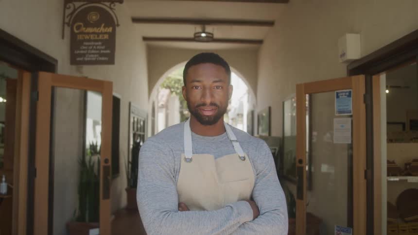 An employee wearing a light colored apron and a grey shirt stands with his arms crossed looking down with a serious expression then looks directly at the camera and smiles