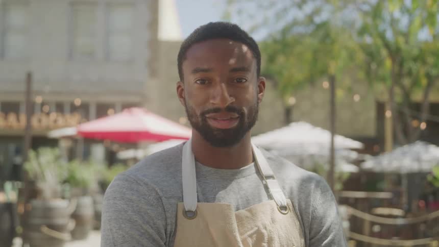 A young adult man with a beard wearing a grey shirt and a light colored apron looks directly at the camera with a subtle smile standing outdoors in what appears to be a cafe or restaurant patio with blurred tables and umbrellas