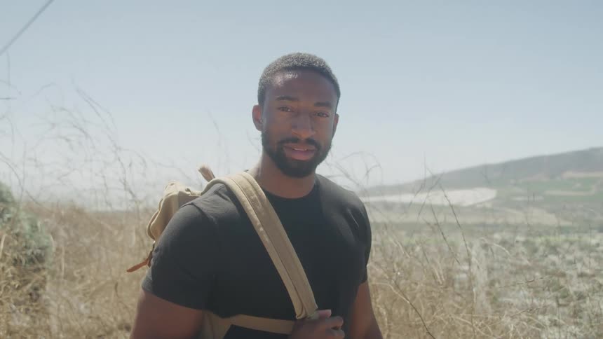 A black man with a backpack over one shoulder in a field turns his head and smiles at the camera