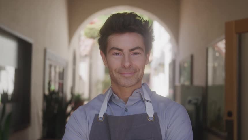 A man with dark hair wearing a light blue collared shirt and a grey apron looks directly at the camera with a friendly smile standing in front of the arched entrance of a building