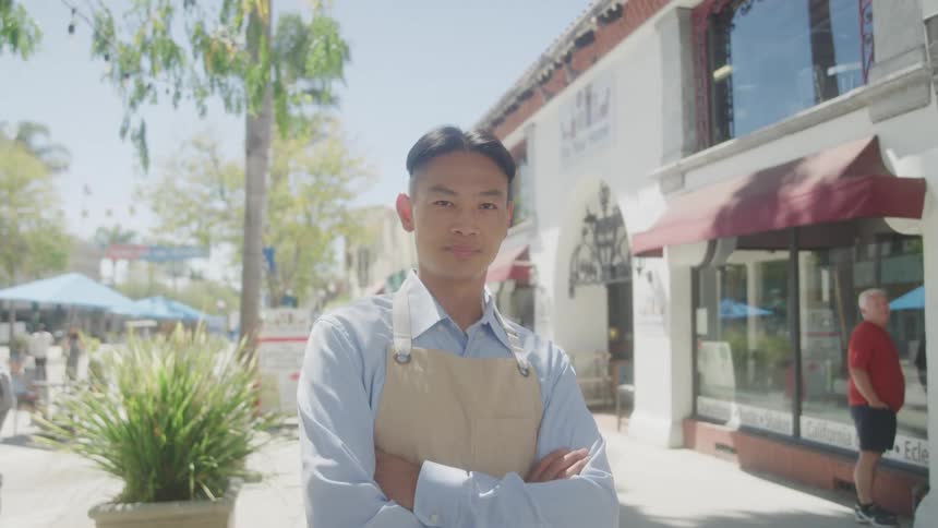 A smiling man with short dark hair and a lightcolored apron stands with his arms crossed on a sunny street looking directly at the camera he is positioned in front of a white building with an awning suggesting a storefront or business