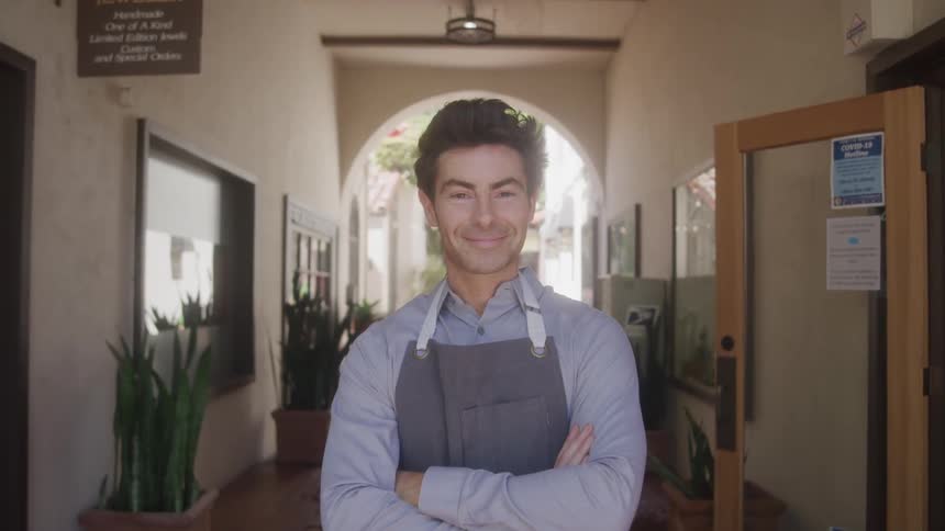 An employee with a subtle smile and his arms crossed wears a grey shirt and a dark apron standing in the arched doorway of a store