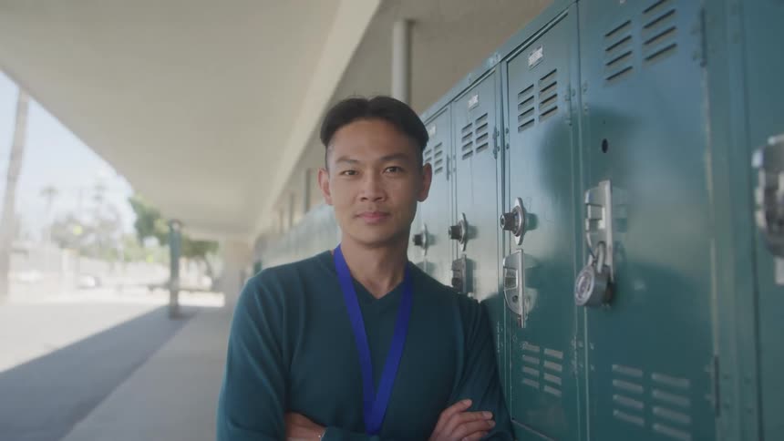 A man stands with his arms crossed and leans against a row of lockers looking directly at the camera he is wearing a blue lanyard around his neck suggesting a school or institutional setting