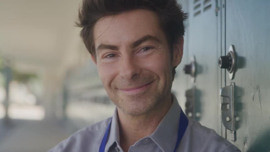 A male teacher with dark styled hair and a blue lanyard is standing in what appears to be a school hallway leaning against green lockers he smiles directly at the camera conveying a friendly and approachable demeanor