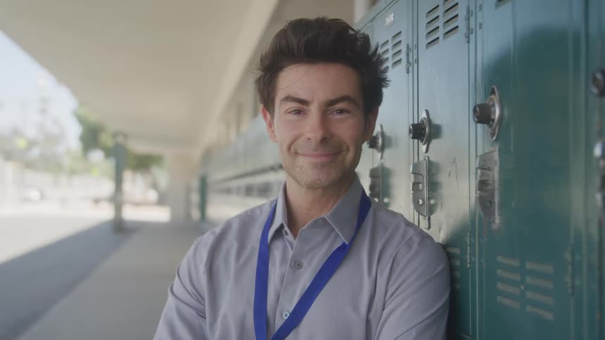 A professionallooking man with a lanyard around his neck standing in a school hallway with a sincere and friendly smile as he leans casually against a row of teal lockers