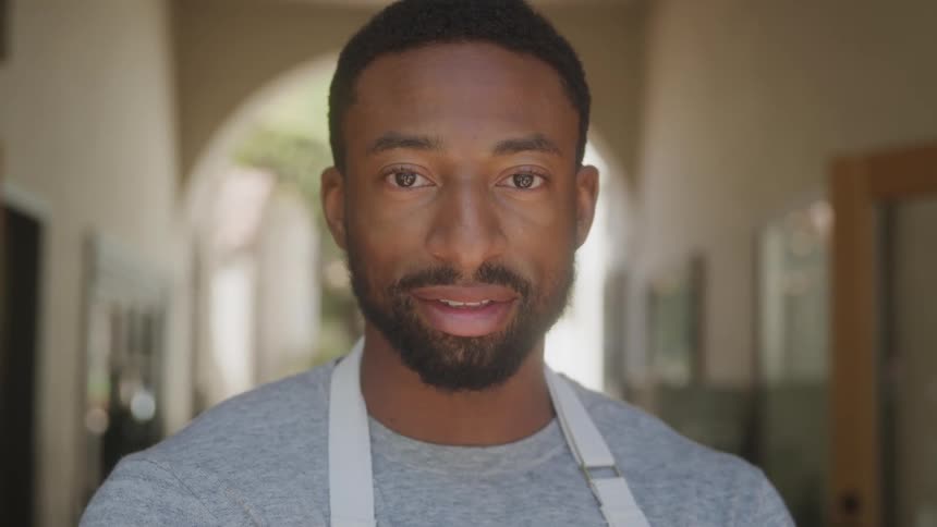 A young adult man with a beard and short hair wearing a grey shirt and a light colored apron looks directly at the camera with a subtle smile in an indoor setting with blurred archways in the background