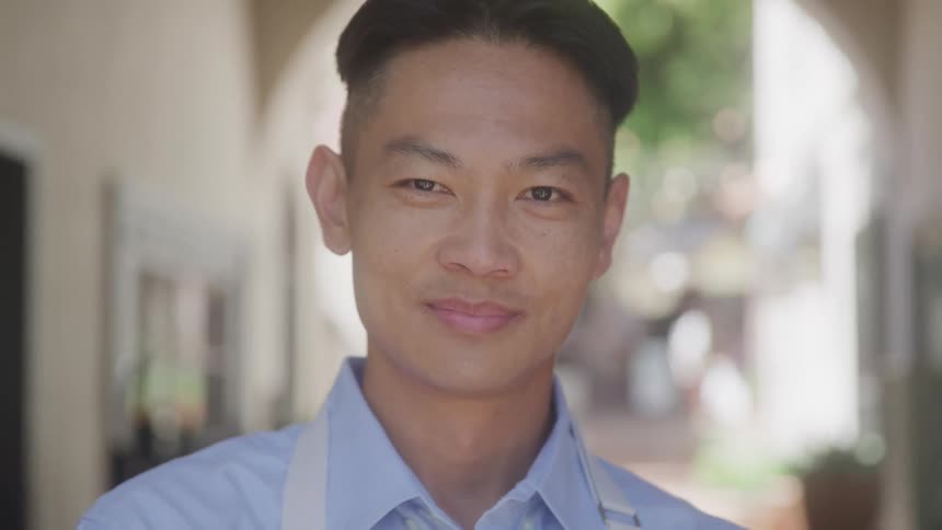 A closeup captures a young man with dark styled hair looking directly at the camera with a gentle smile he is wearing a light blue shirt with a lighter blue apron or strap over his shoulder suggesting a professional or serviceoriented setting
