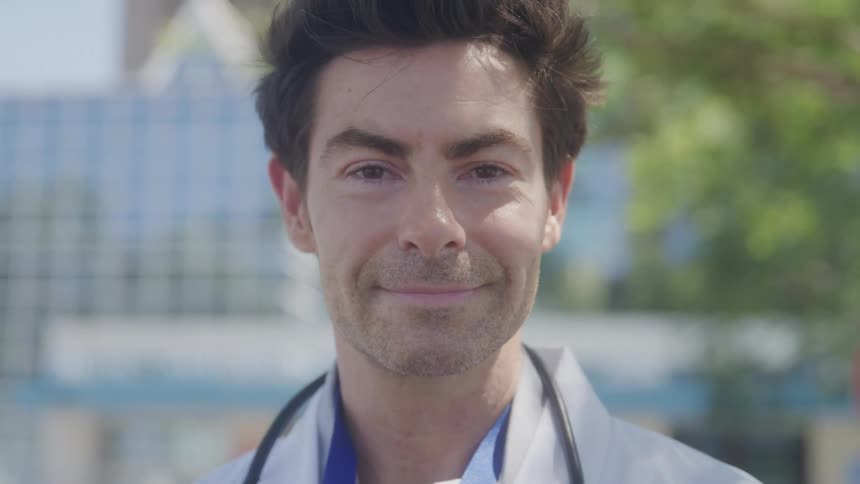 A doctor wearing a stethoscope around his neck is looking away from the camera and then turns to look at the camera he smiles softly and the scene ends with him looking directly into the camera