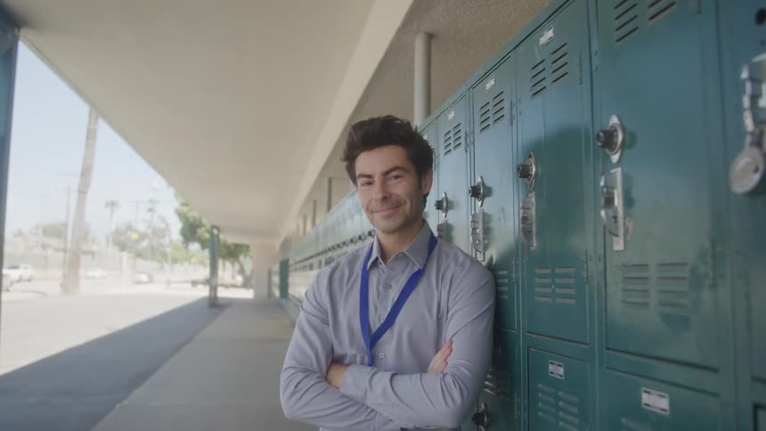 A male teacher possibly in a school hallway is standing with his arms crossed leaning against green lockers he is looking off to the side with a pensive expression and then turns to smile directly at the camera