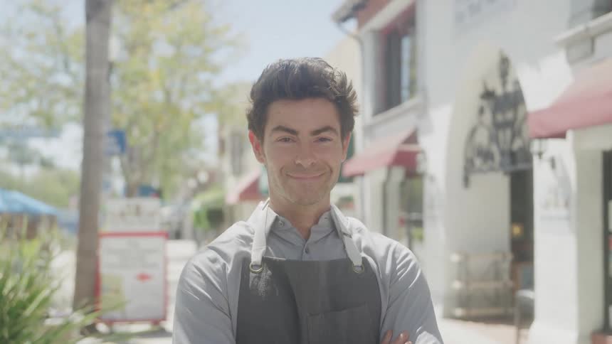 A young adult man wearing a light colored collared shirt and a dark apron stands with his arms crossed and smiles directly at the camera with a bright outdoor street and buildings in the background