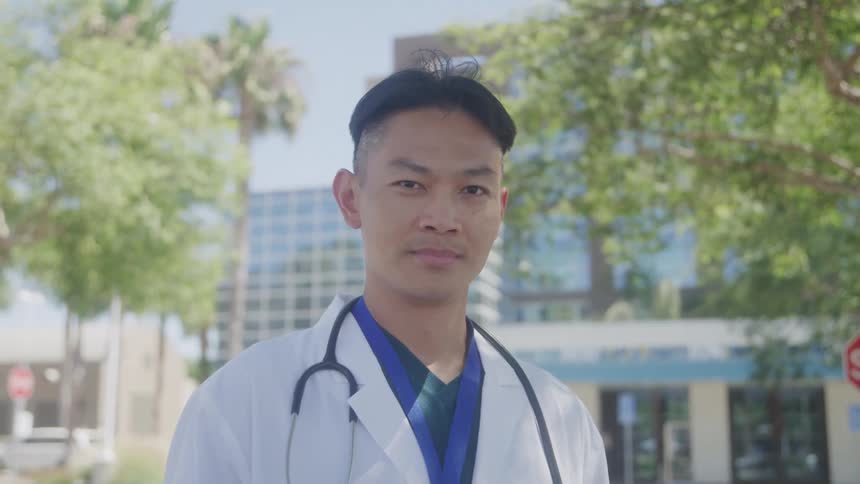 A doctor stands outdoors in front of a modern medical building wearing a white coat and stethoscope with a composed expression