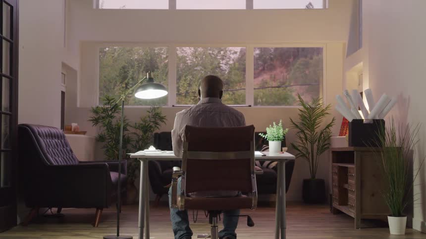 A man sits indoors behind a desk in a home office with a reading lamp on the side of the desk