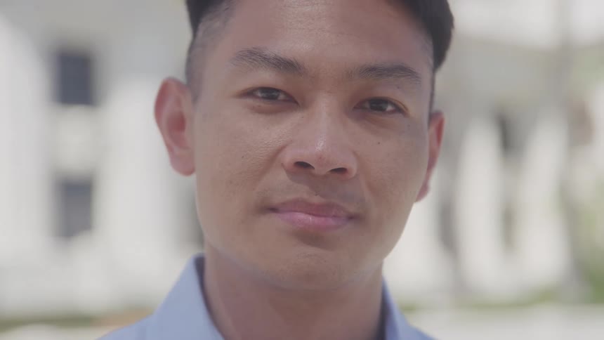 A closeup captures a young man with a slight smile looking directly at the camera he is wearing a light blue collared shirt and the background is blurred with hints of a lightcolored building