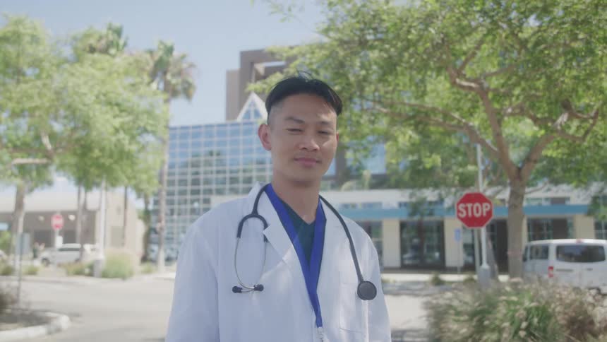 A doctor wearing a stethoscope over his shoulder stands outside a hospital at midday