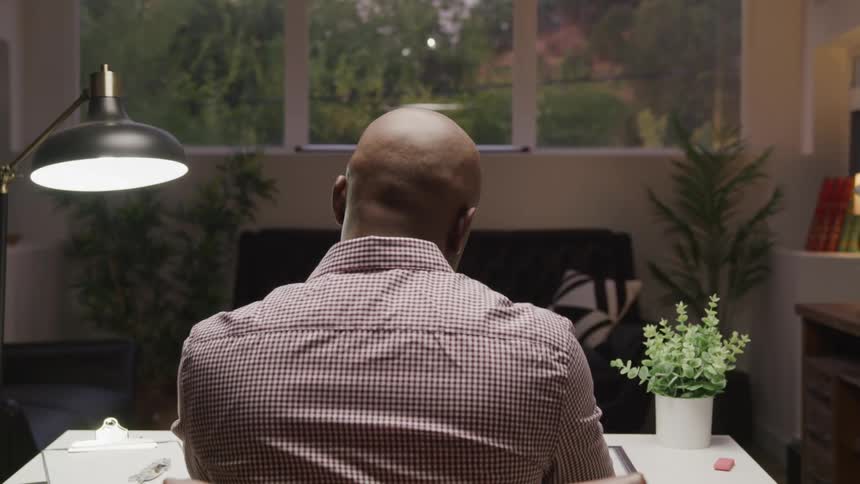 A bald man in a plaid shirt sits with his back to the camera looking out a large window at a lush green landscape a desk lamp and potted plants are visible on the desk in front of him