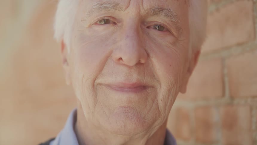Close up portrait of an elderly man standing beside a brick wall outdoors during daytime
