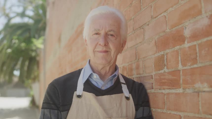 An elderly man with gray hair rests beside a brick wall at midday