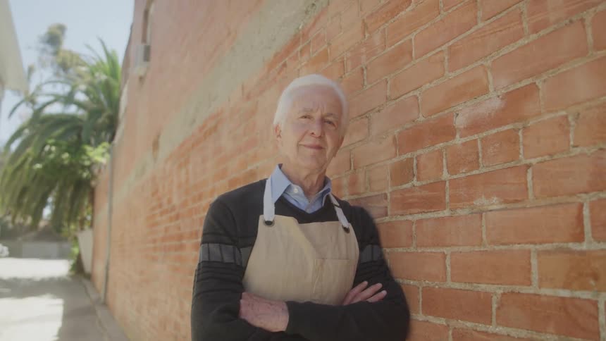 An elderly man with gray hair wearing an apron rests against a brick wall at midday