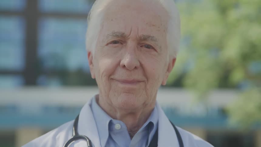 Elderly white haired doctor stands outside of a building on a summer day
