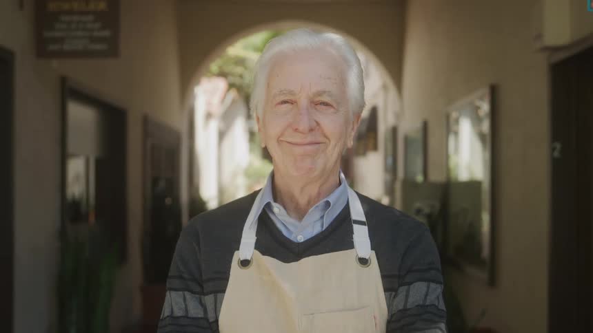 An elderly man smiles warmly while standing in an outdoor corridor