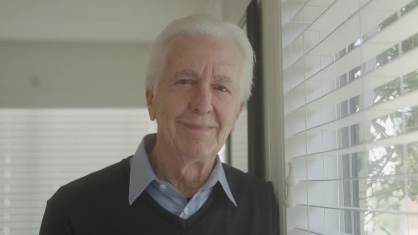 Close up portrait of an elderly man leaning by the window while standing in a room during daytime