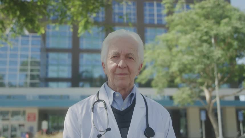Close up portrait of an elderly male doctor standing outdoors in front of a hospital building during daytime