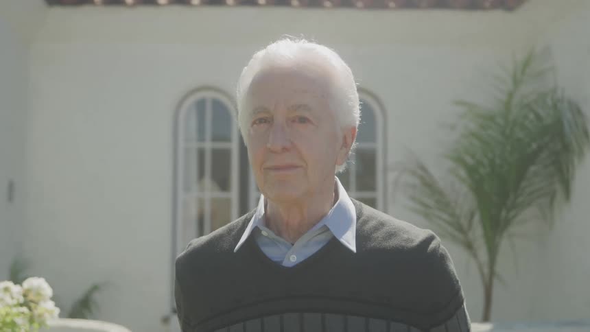 Elderly man standing near an elegant white home looks forward with a smile