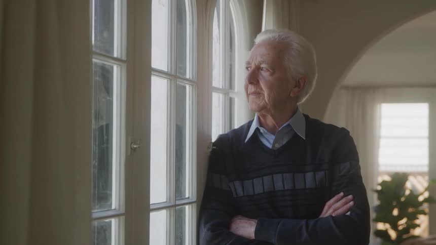 An elderly gentleman with white hair wearing a striped sweater over a collared shirt stands with his arms crossed looking thoughtfully out a large window in a brightly lit room