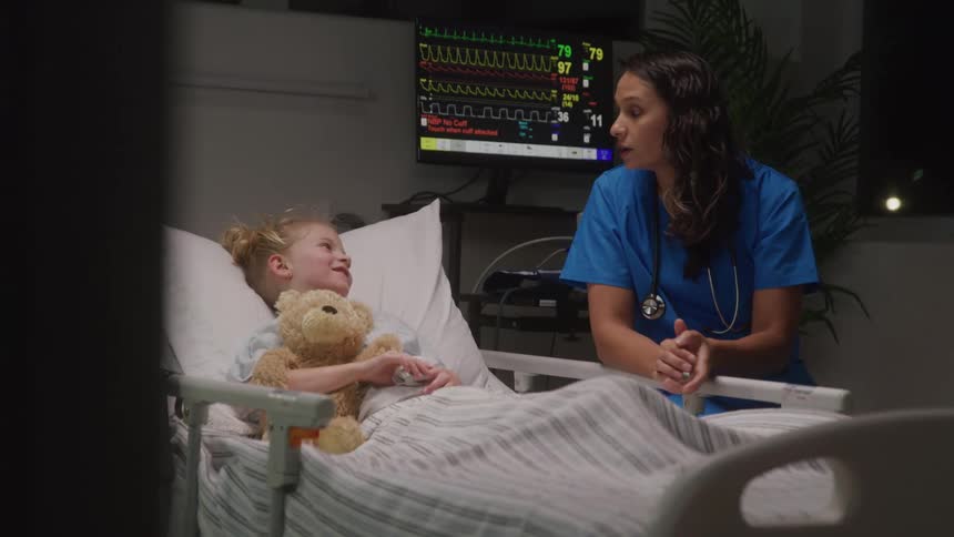 A young adult female nurse in blue scrubs leans over a hospital bed talking to a young girl who is holding a teddy bear with a medical monitor displaying vital signs and a plant in the background