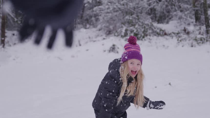 A young girl with blond hair wearing a purple and orange knitted hat and a black winter coat playfully throws snow into the air laughing as she engages in the activity in a snowy forest