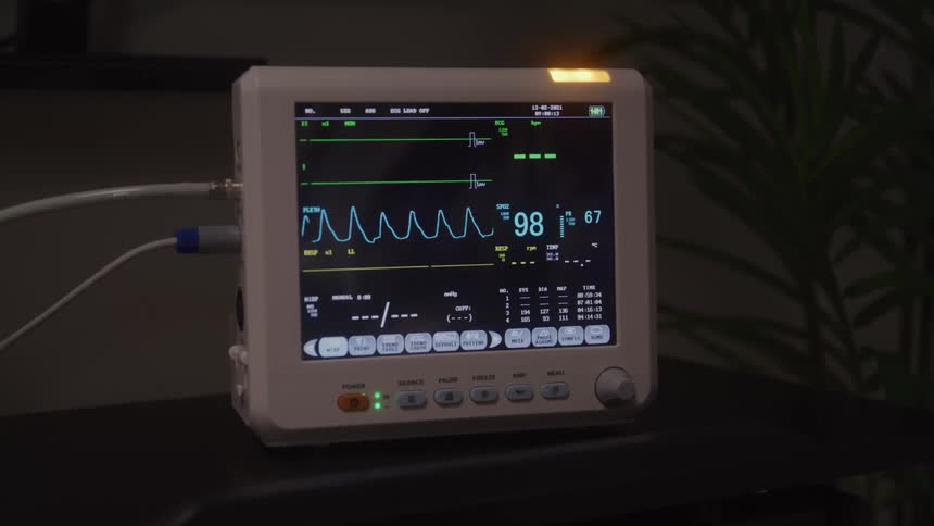 A close view of a medical monitor shows a green waveform and other vital sign data with cables attached to the device