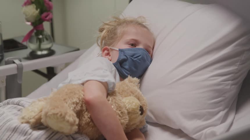 Little girl wearing face mask hugging a teddy bear laying in a hospital bed
