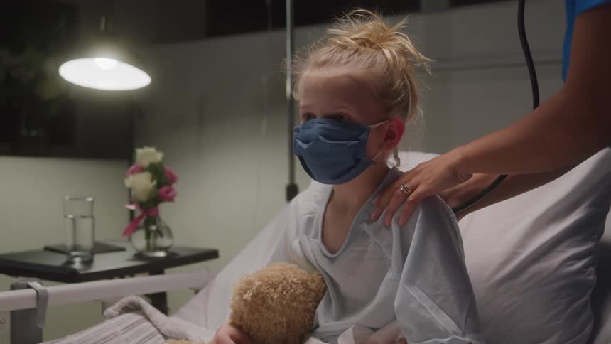 A young girl with blond hair wearing a blue face mask and holding a teddy bear sits up in a hospital bed as a nurse places a stethoscope on her back during an examination