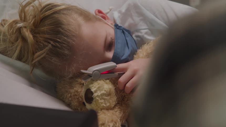 A young child with light colored hair wearing a blue face mask and holding a brown teddy bear lies sleeping in a hospital bed with a pulse oximeter attached to their finger