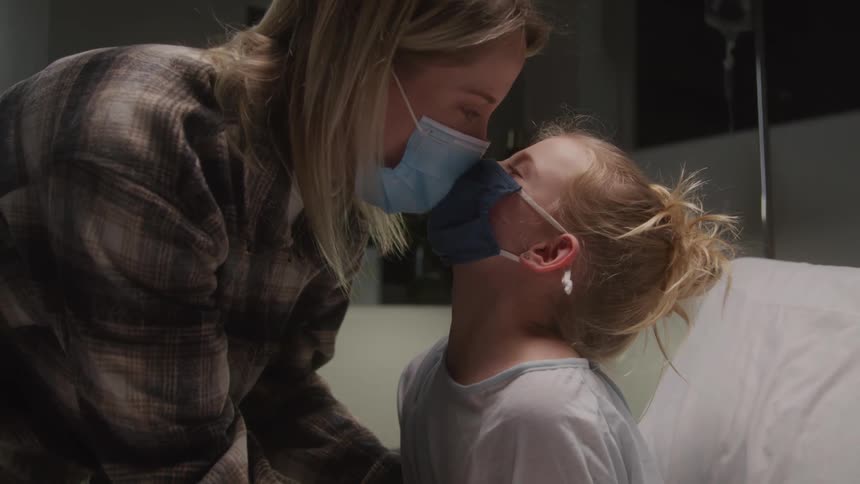 A kid sits in a hospital bed with her mother beside her both wearing face masks and kissing each other