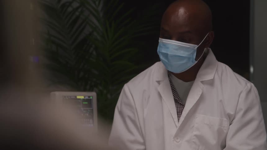 A medical professional a man with a bald head wears a blue face mask and a white lab coat looking forward with a serious expression with medical equipment displaying vital signs and a green plant in the background