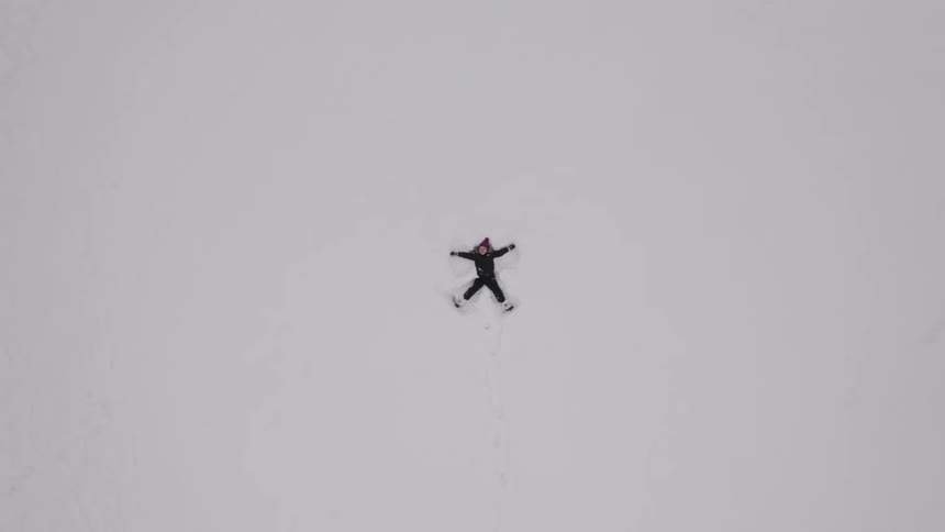 Aerial of a child in a green snowsuit making a snow angel in the snow