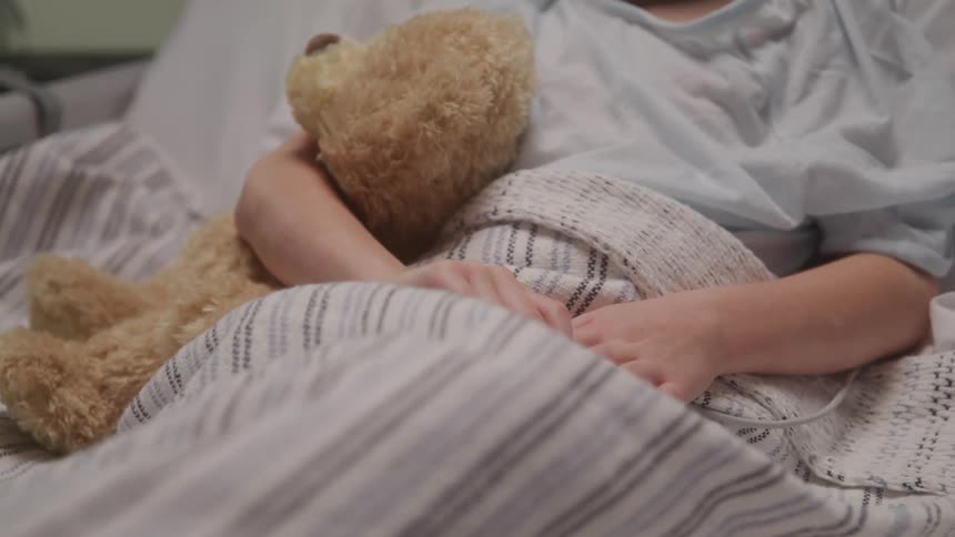 A young child lies in a hospital bed holding a brown teddy bear and a medical device attached to their finger under white striped blankets