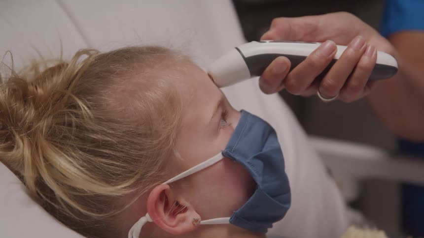 A young girl with blond hair wearing a blue face mask lies on a white pillow as a medical professional likely a nurse uses a white and black digital thermometer to check her temperature