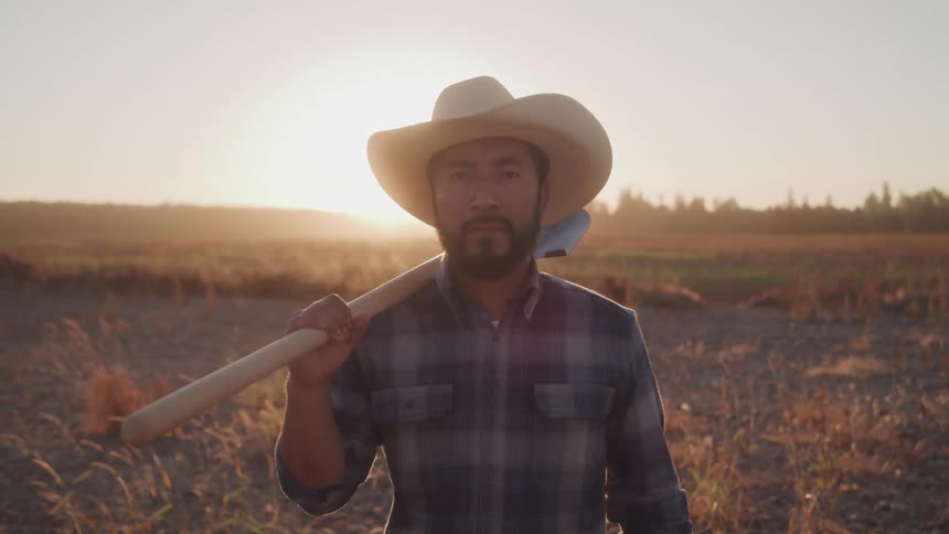 A farmer stands in a field with the orange setting sun in sight holding a shovel on his shoulder and wearing a sunhat