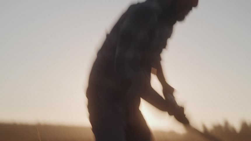 A male farmer in a hat is silhouetted against a bright sunset sky swinging a pickaxe vigorously while working in a field