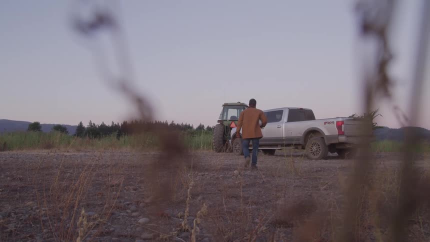 A lowangle wide shot shows a person walking across a vast dry field towards a parked pickup truck and a large piece of farm machinery with trees and hills in the distant background