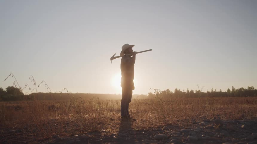 A male farmer carries a pick axe over his shoulder while standing in a field outdoors at sunset