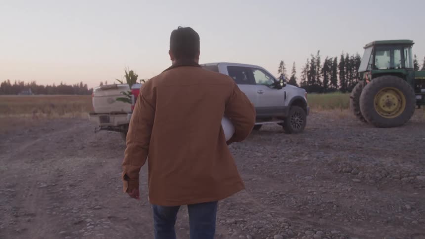 A man wearing a brown jacket holds a hard hat and walks toward his truck in a field with a tractor parked beside it at midday