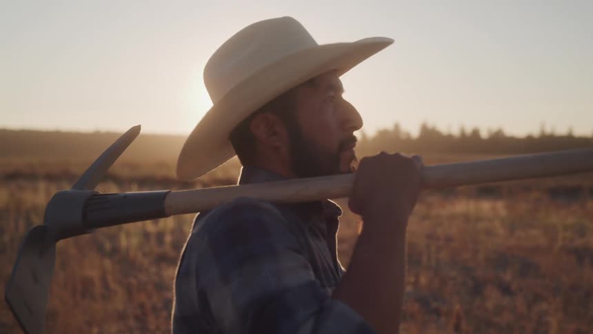 A male farmer with a beard wearing a light colored cowboy hat and a plaid shirt is seen in profile carrying a pickaxe over his shoulder looking out at a field during sunset with bright sun flare