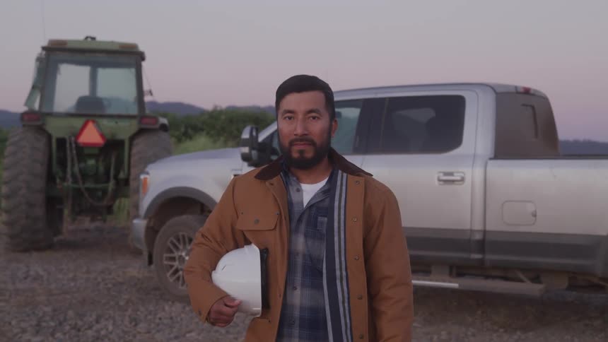 Rancher holding a hardhat standing in front of truck and tractor on a farm