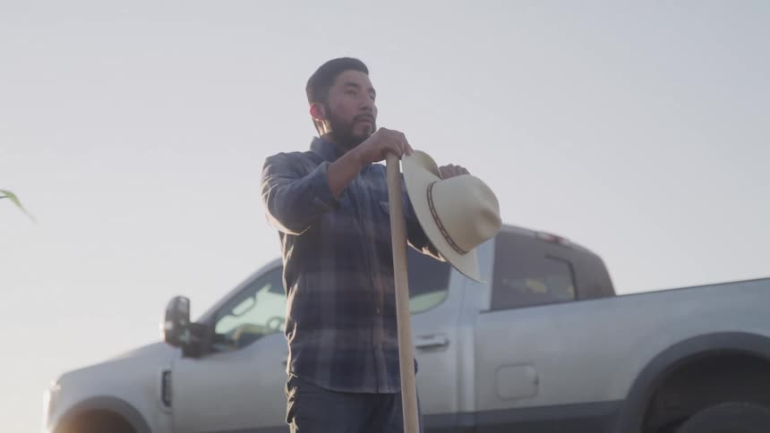 Man in a hat on a farm weeding and a white pickup truck in the background