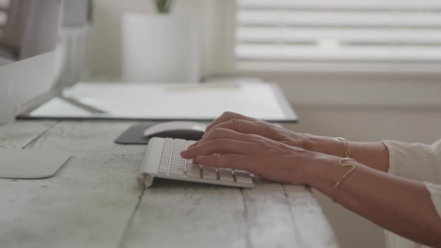 A close view of an adult persons hands wearing light colored bracelets typing on a white computer keyboard with a mouse and monitor on a light colored desk in the background
